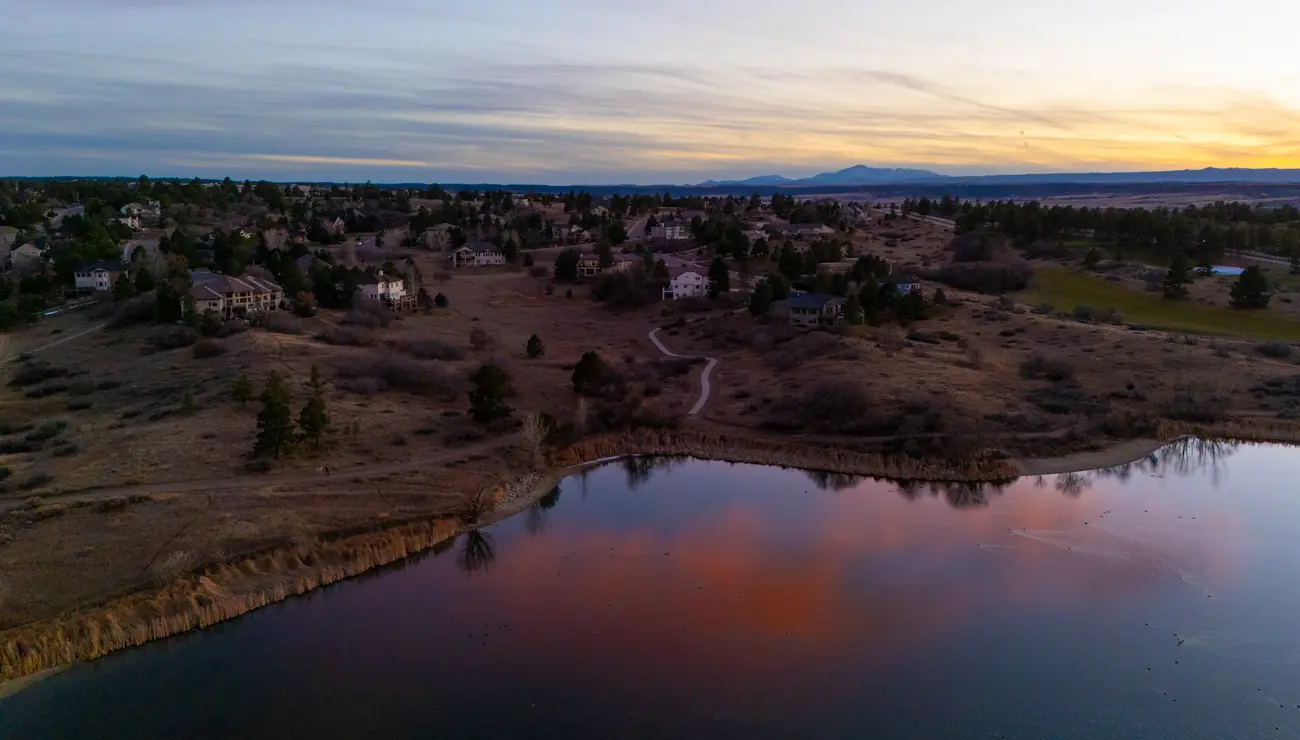 The lake at dusk holding the reflection of a pink cloud, a trail winding up toward homes on the ridge.